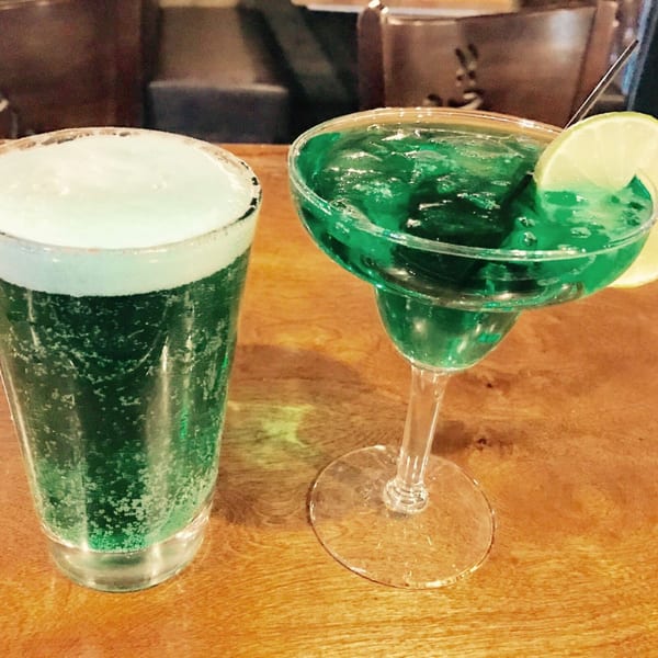 Two green drinks, one foamy beer and one margarita with a lime, on a wooden table.