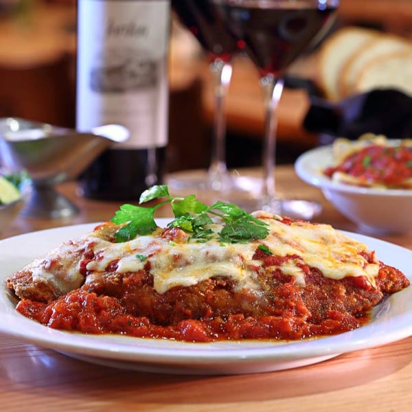 Chicken Parmesan with melted cheese and red sauce, garnished with parsley, alongside wine and pasta.